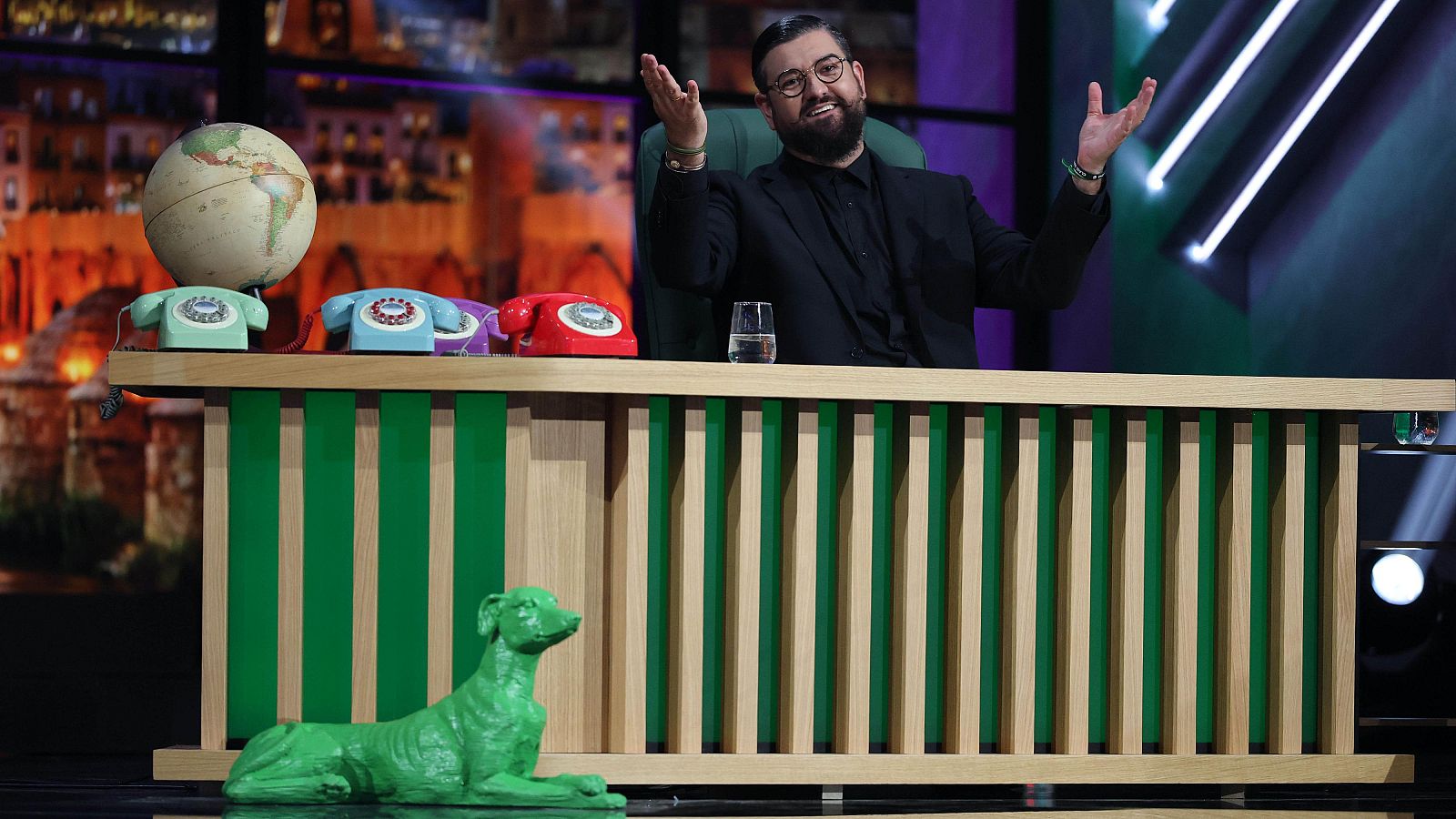 Manu Sánchez, con gafas y barba, sonríe gesticulando detrás de un escritorio con un globo terráqueo, teléfonos de disco y un perro verde en primer plano, en un estudio de televisión.