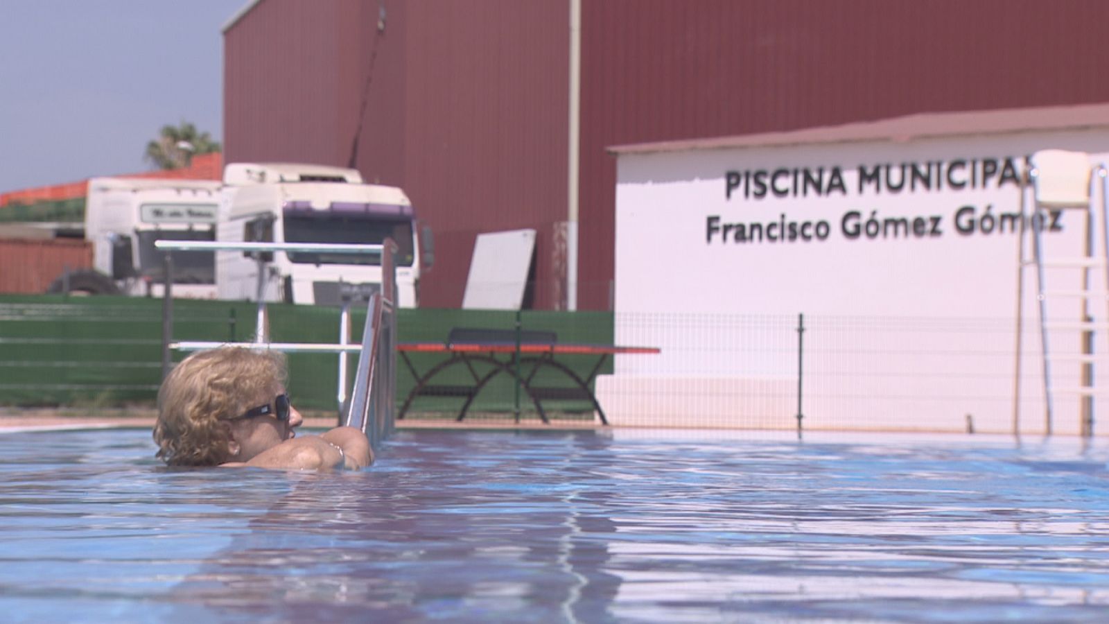 Mujer mayor con gafas de sol se baña en una piscina municipal, apoyada en la escalera. Al fondo, el nombre "PISCINA MUNICIPAL Francisco Gómez Gómez".