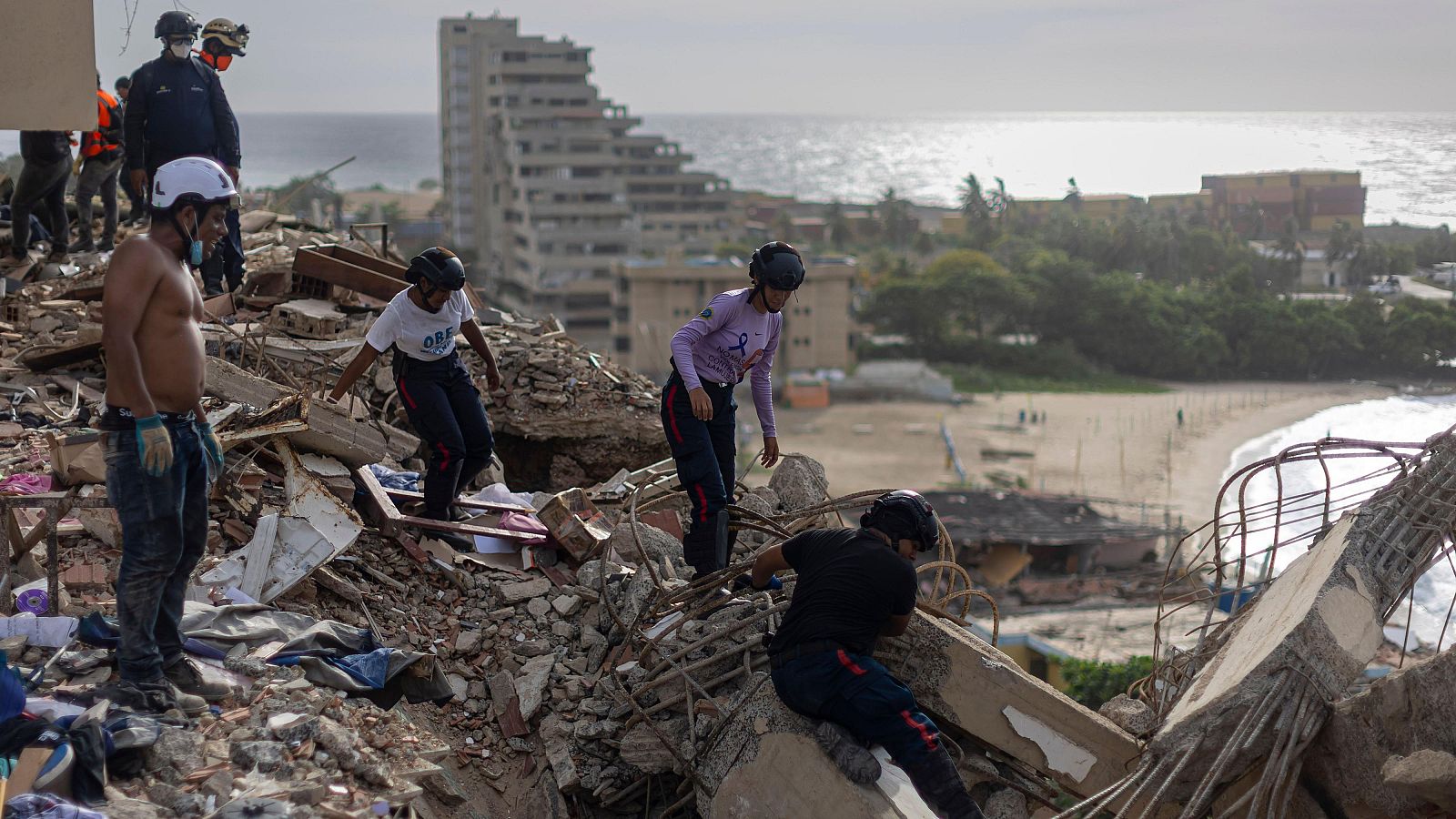 Rescatistas buscan entre escombros en una zona costera devastada, con el mar y vegetación al fondo. Varios equipos trabajan activamente en la remoción de escombros.