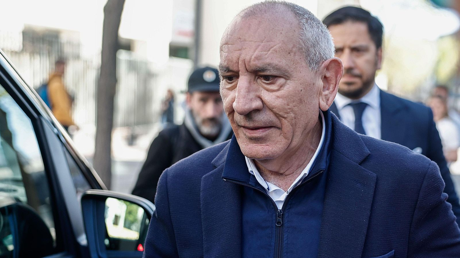 Primer plano de José Ángel González, ex DAO de la Policía Nacional, con expresión seria. Viste chaqueta azul marino y camisa blanca. Al fondo, otras dos personas y un vehículo.
