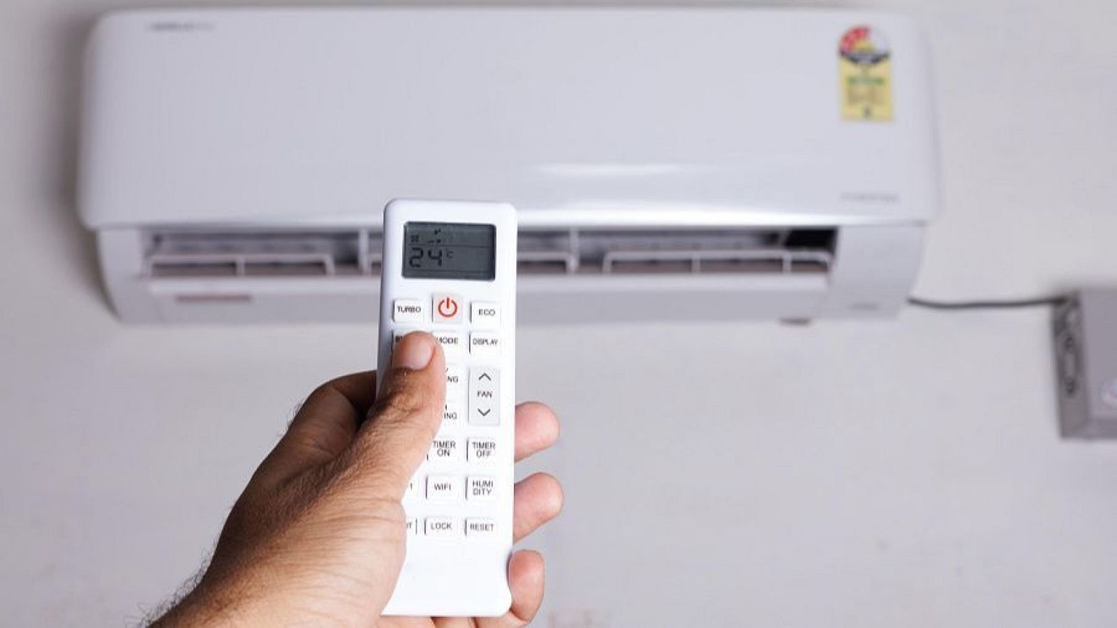 Mano operando mando de aire acondicionado con pantalla a 24°C, mostrando botones como TURBO, ECO, MODE y TIMER ON. Unidad split blanca visible al fondo.