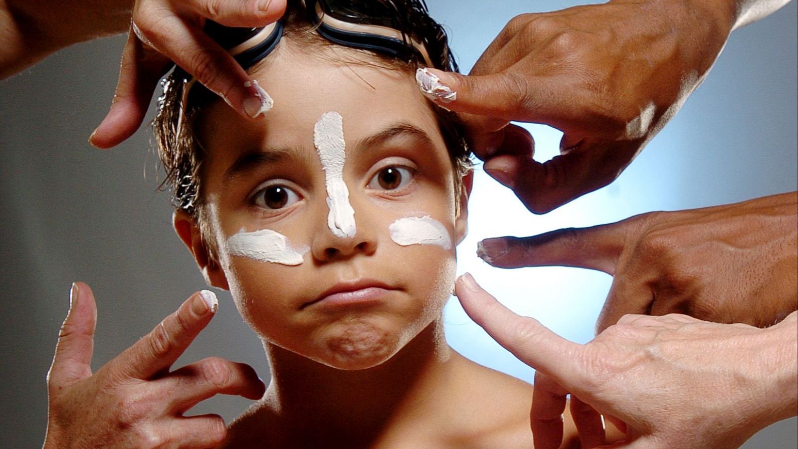 Niño con gafas de natación y crema blanca en el rostro, rodeado por manos de adultos que se la aplican. Fondo degradado y luz suave.