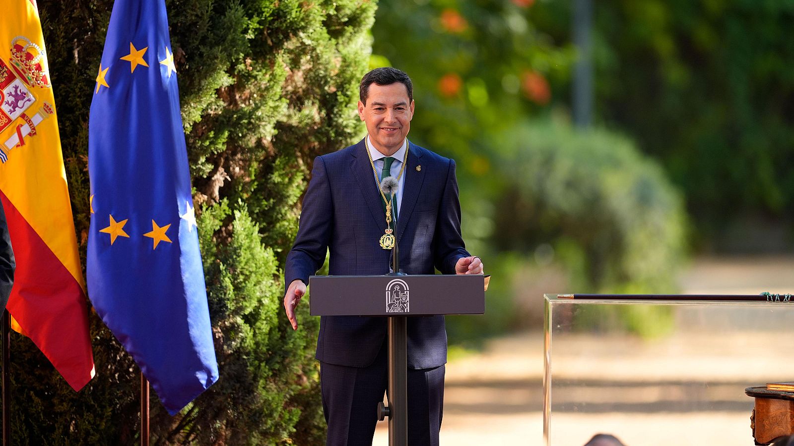 Juan Manuel Moreno, presidente de la Junta de Andalucía, sonríe con un traje azul y corbata verde, luciendo un collar ceremonial dorado, con las banderas de España y la UE al fondo.