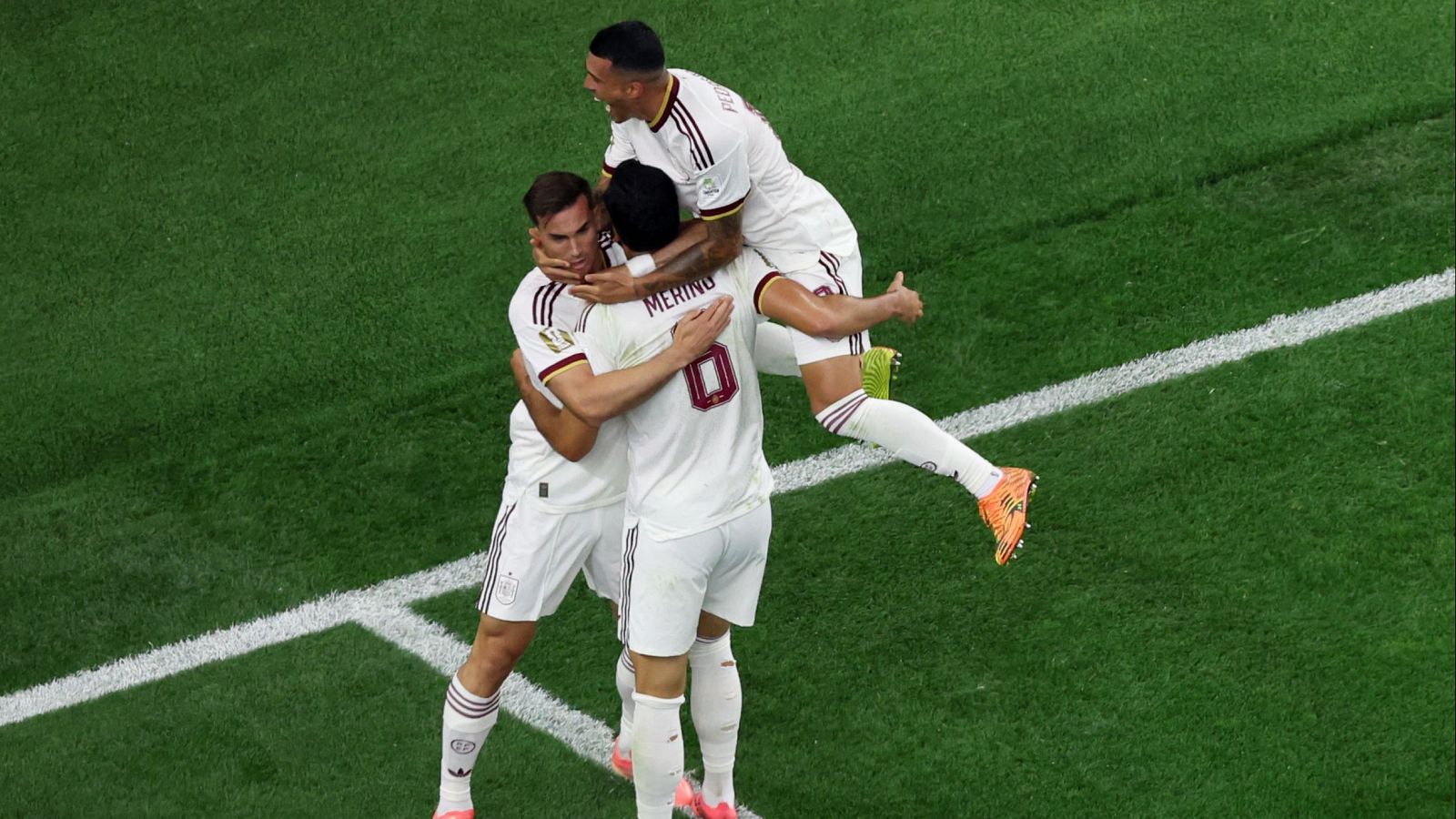 Futbolistas de España celebran un gol. Se distingue a Merino (8) abrazado por compañeros, uno de ellos salta con el pulgar arriba. Botas naranjas visibles.