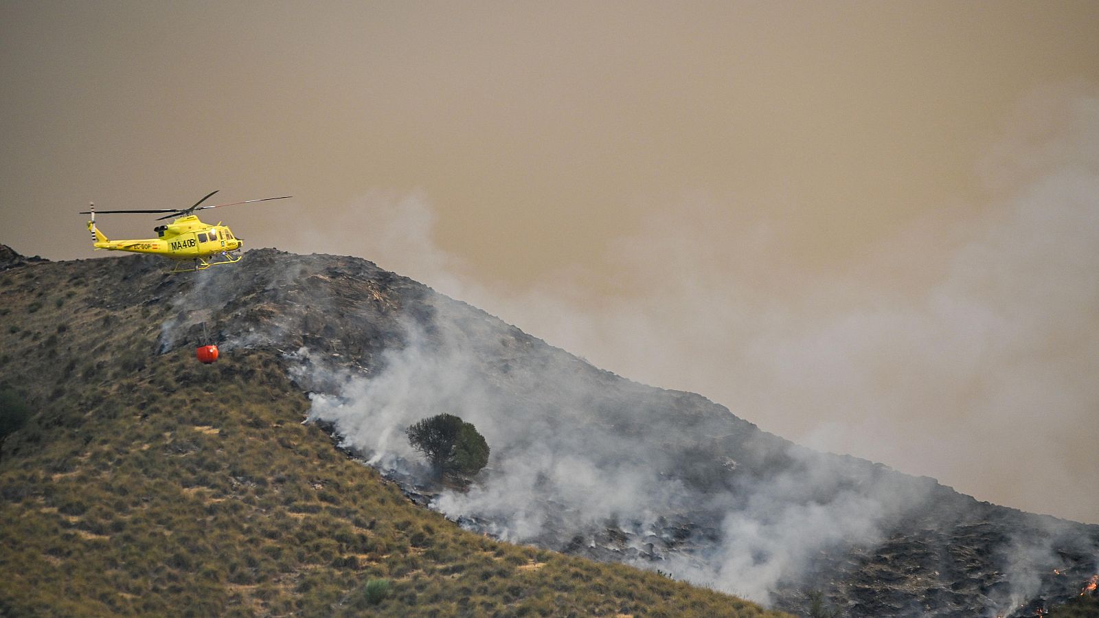 Helicóptero amarillo con cubo rojo combate incendio forestal en ladera montañosa. Humo denso cubre el cielo de tono marrón.