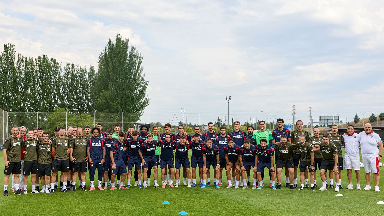 Fotografía de grupo de un equipo de fútbol con 40-50 hombres en un campo de césped, vistiendo uniformes azul marino, verde oliva y polos blancos, con el logo "KOSNER" visible.