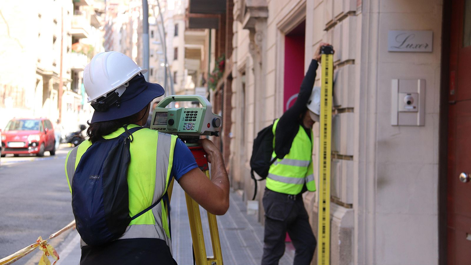 Técnicos realizan mediciones topográficas en una calle urbana, uno con estación total y otro con mira, frente a un edificio con cartel "Luxor".