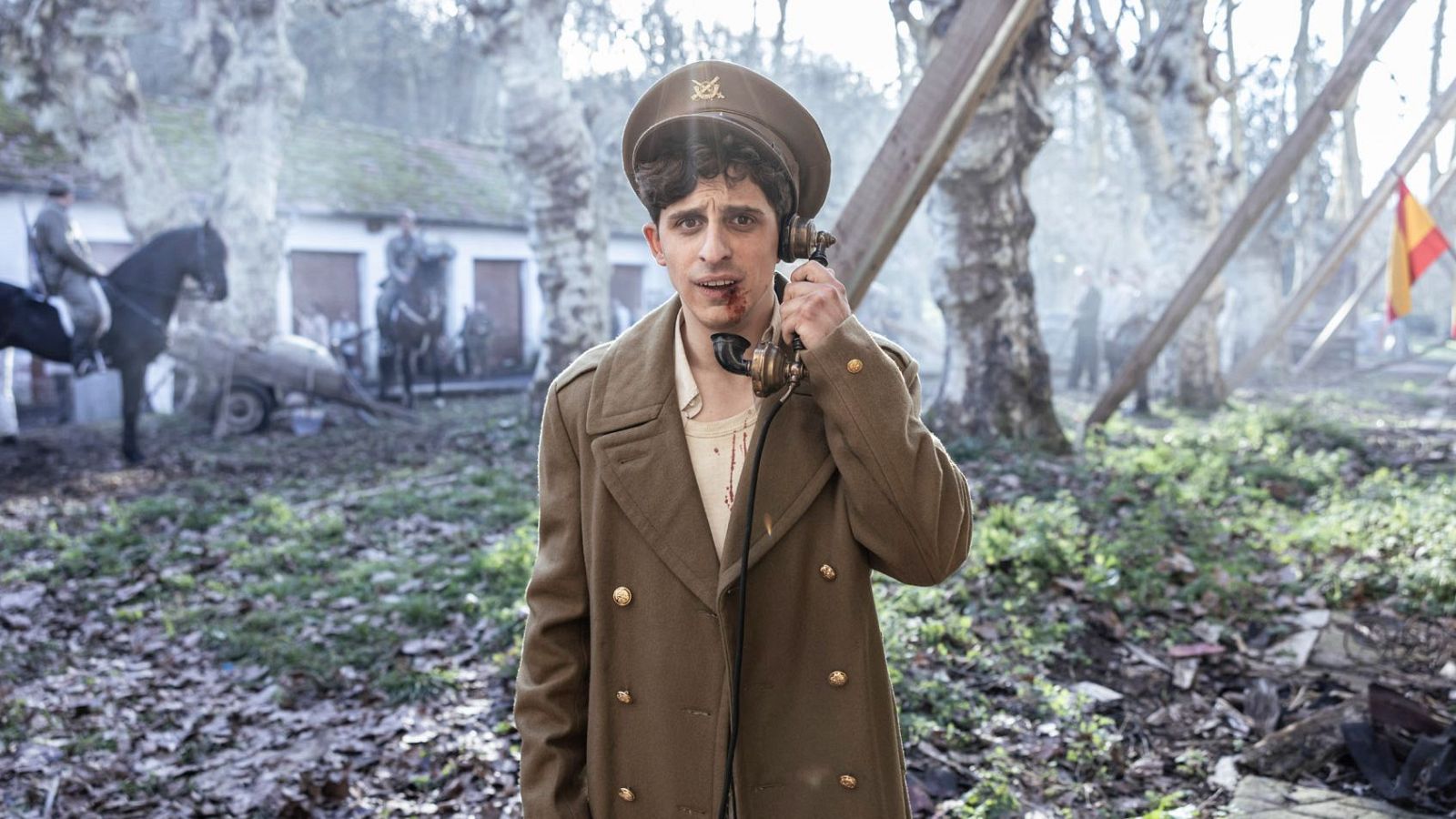 David Verdaguer como Miguel Gila, herido y con un teléfono antiguo, en un dramático escenario de guerra con soldados y una bandera española.