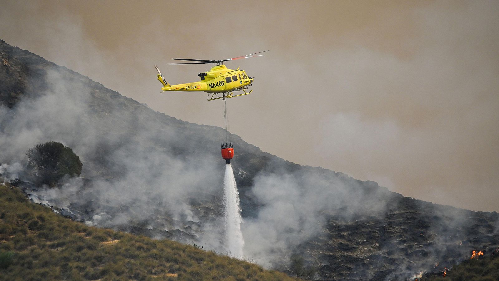 Helicóptero MA-408 con bandera de España arroja agua sobre incendio forestal en ladera montañosa con humo y llamas activas.