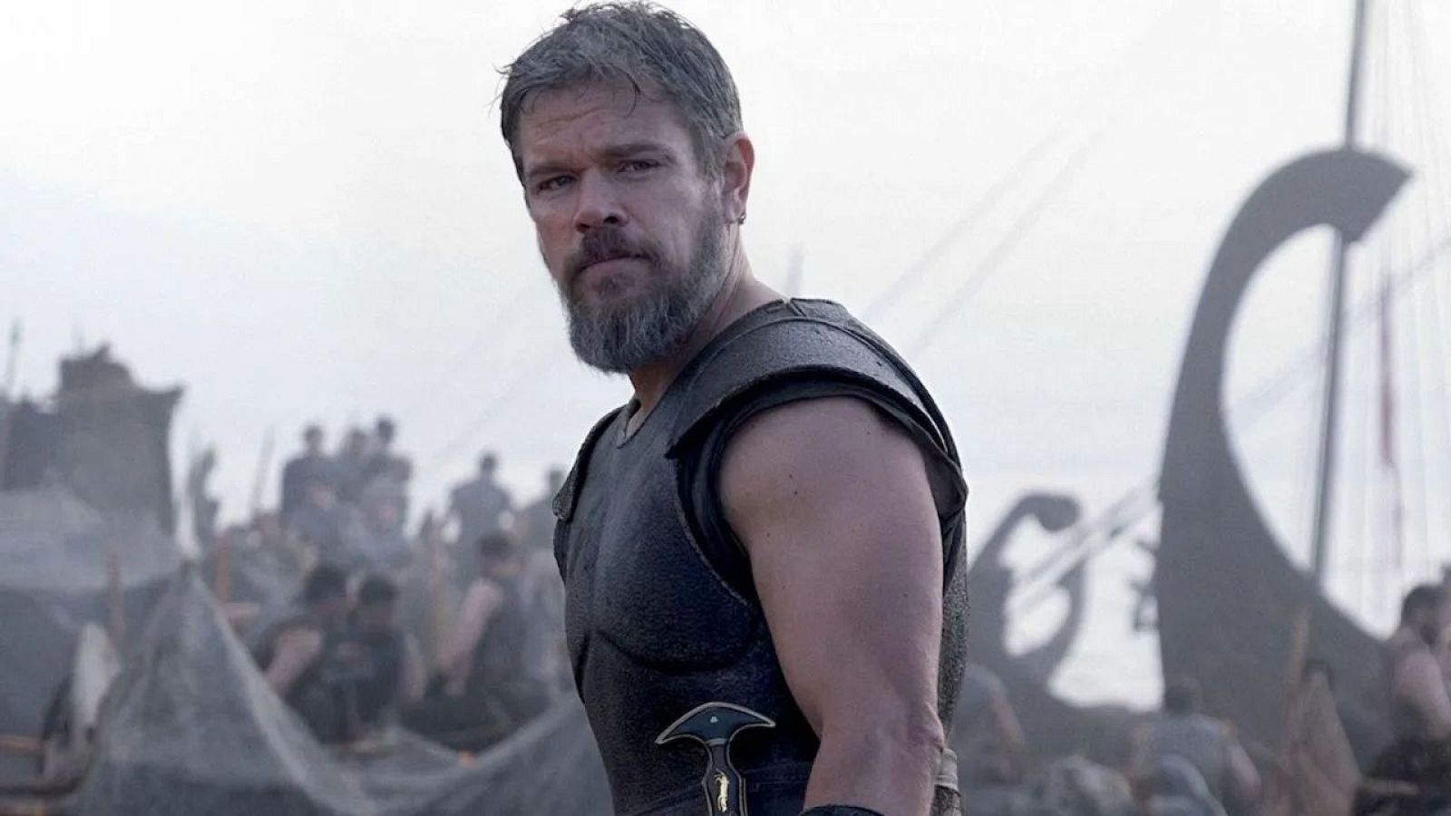 Matt Damon, con armadura de cuero oscura y barba canosa, aparece en primer plano en un fotograma de 'La Odisea', con un campamento vikingo y barcos al fondo.