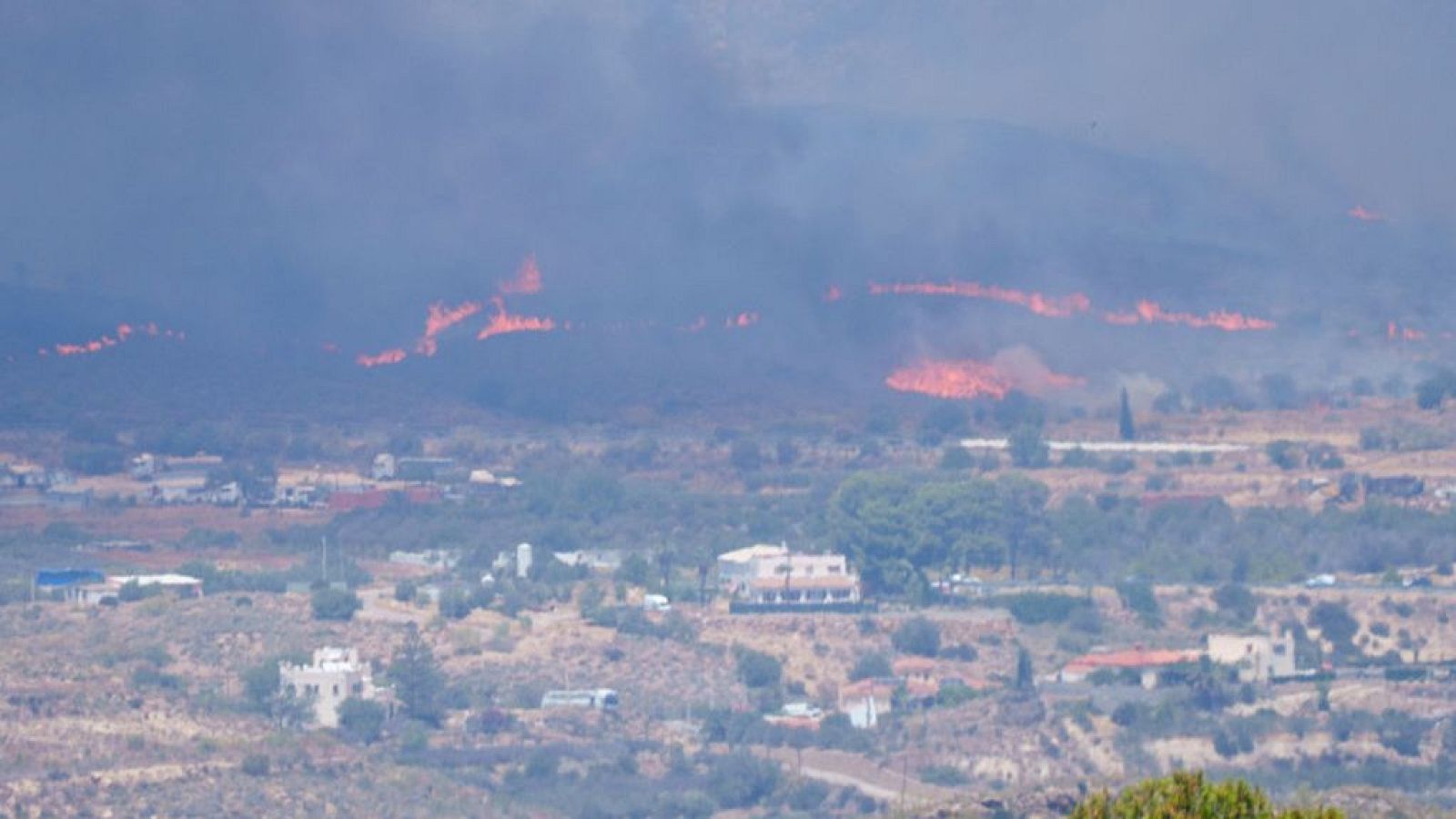 Incendio forestal en Los Gallardos, Almería. Llamas y humo cubren la ladera de una montaña, con casas blancas y edificios en el plano medio, transmitiendo una sensación de peligro.