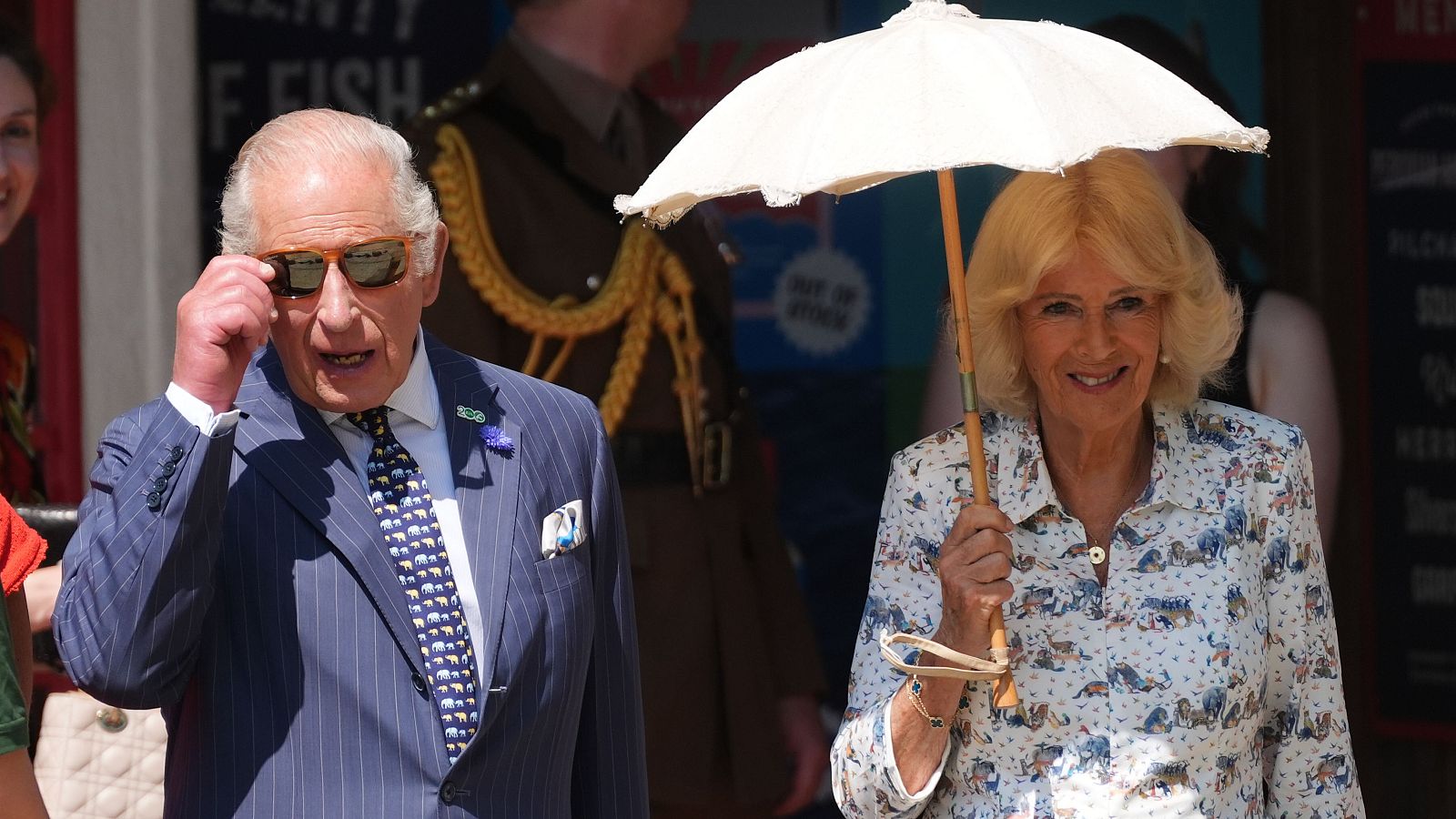 El Rey Carlos III y la Reina Camila en un evento soleado. Él viste traje y ajusta sus gafas de sol, ella lleva blusa estampada y parasol.