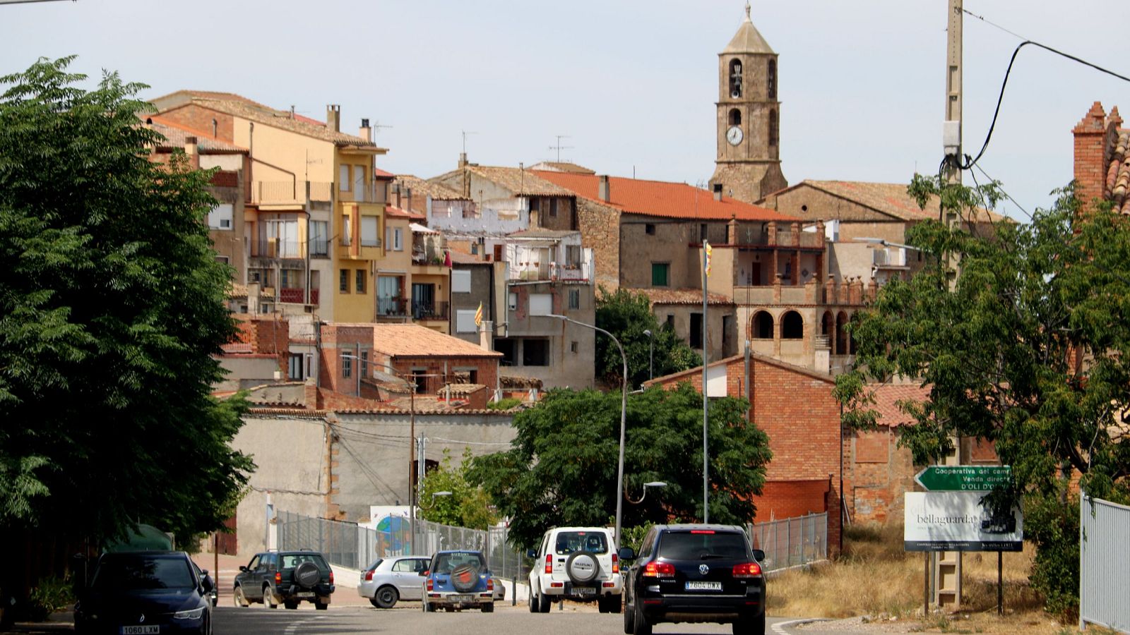 Vista de la entrada a Bellaguarda con carretera, vehículos, edificios tradicionales y un campanario de piedra con reloj al fondo.
