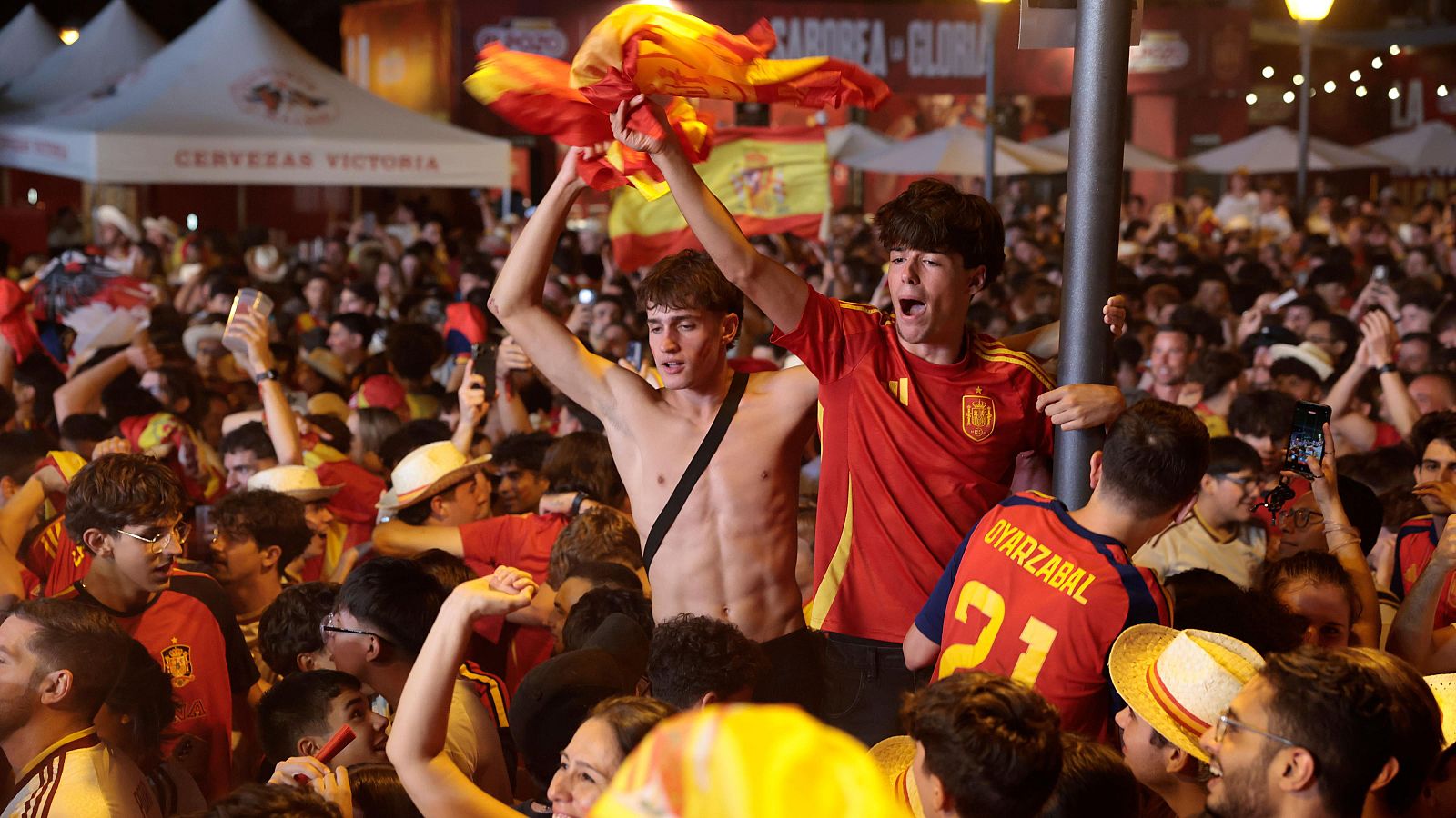 Aficionados españoles celebran eufóricamente, ondeando banderas y vistiendo camisetas rojas y amarillas. Dos jóvenes son el foco, uno con el torso desnudo y otro con la camiseta de Oyarzabal, en una plaza iluminada.