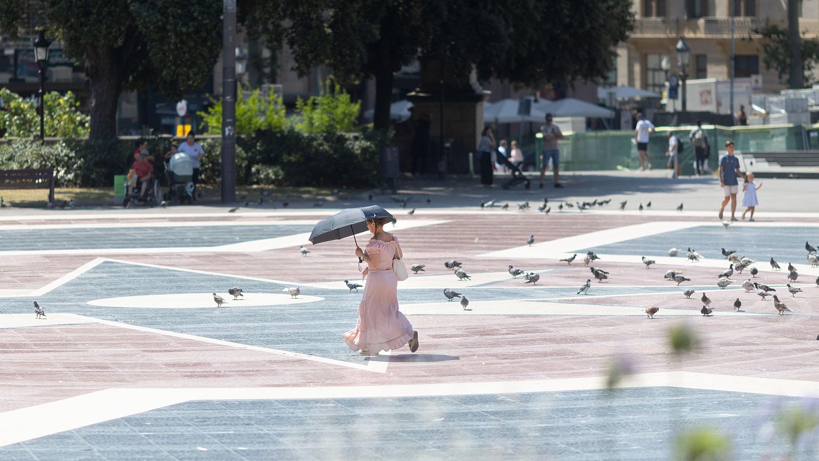 Mujer con paraguas oscuro se protege del sol en una plaza con suelo geométrico y palomas. Al fondo, árboles, bancos y edificios urbanos.