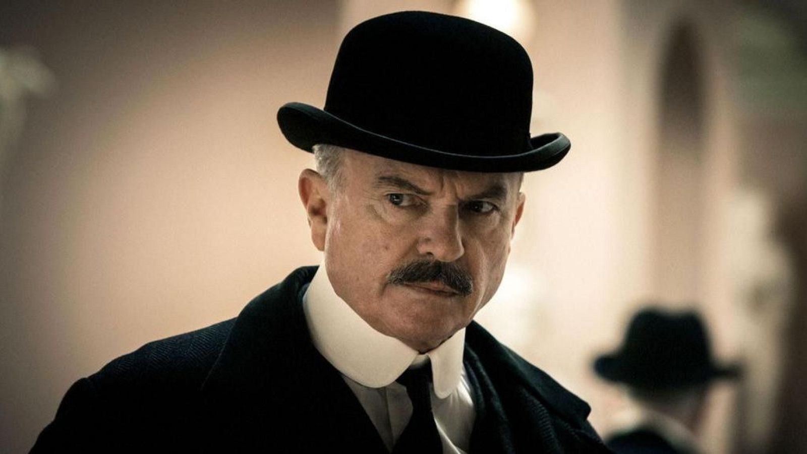 Sam Neill, con sombrero bombín y expresión seria, en un primer plano. Fondo desenfocado con silueta de otra persona.