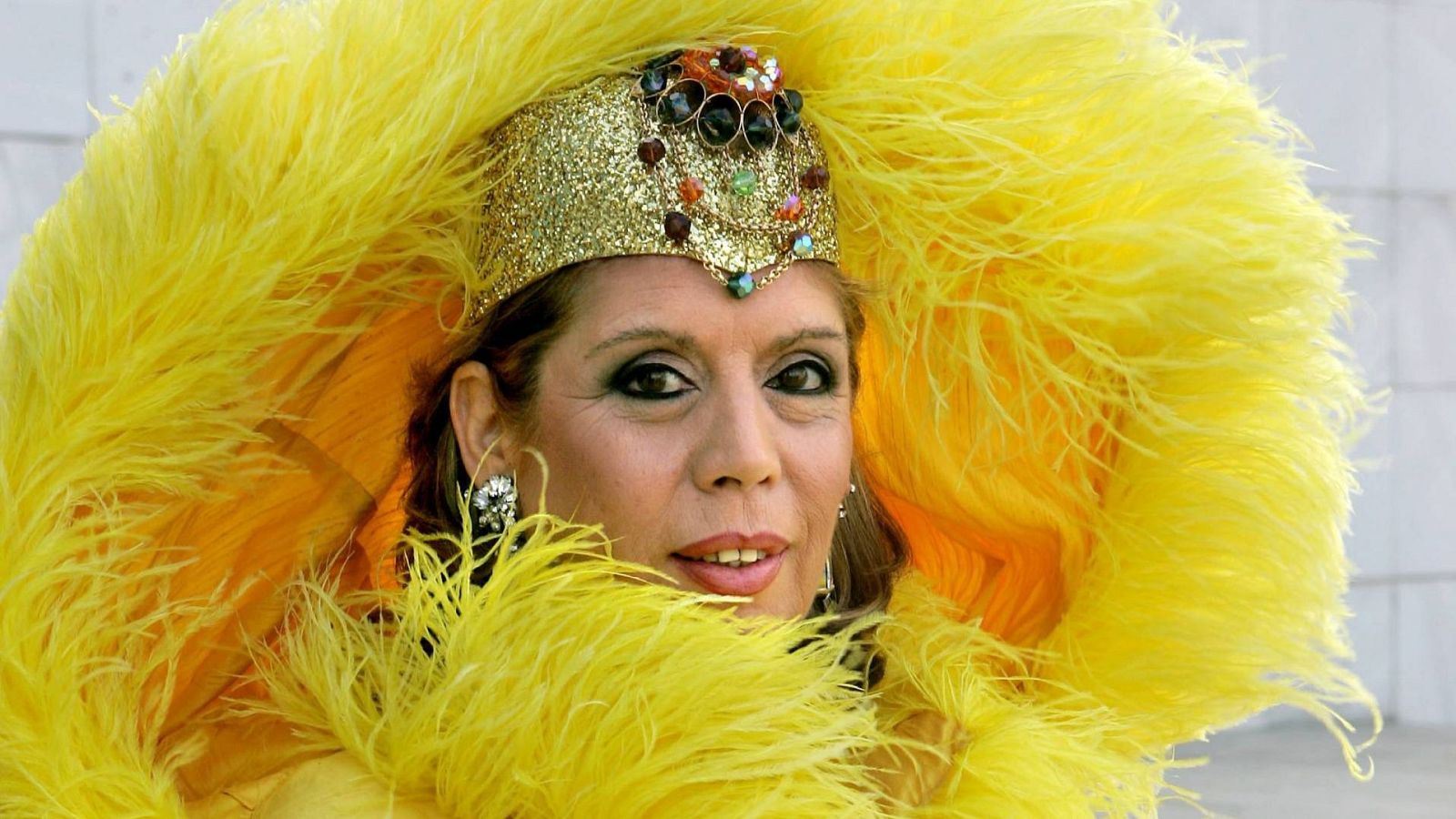 Primer plano de María Jiménez con un espectacular tocado de plumas amarillas, corona dorada con joyas y maquillaje de ojos marcado. Fondo desenfocado.