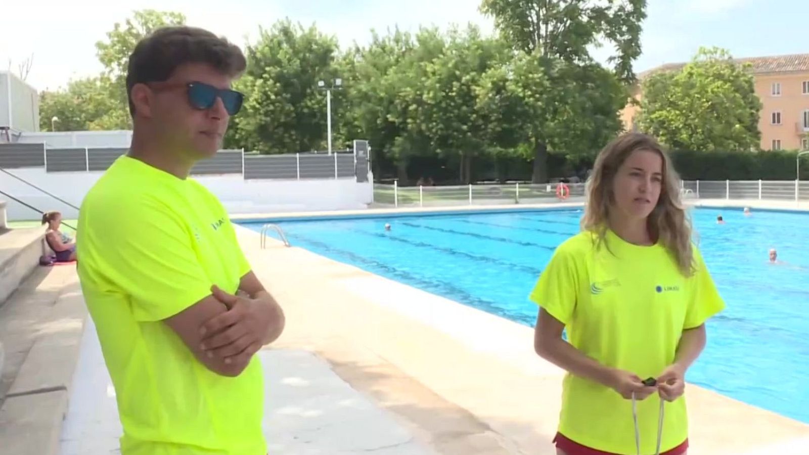 Dos socorristas con camisetas amarillas flúor y gafas de sol rojas posan junto a una piscina azul. La mujer lleva un silbato. Al fondo, bañistas y vegetación.