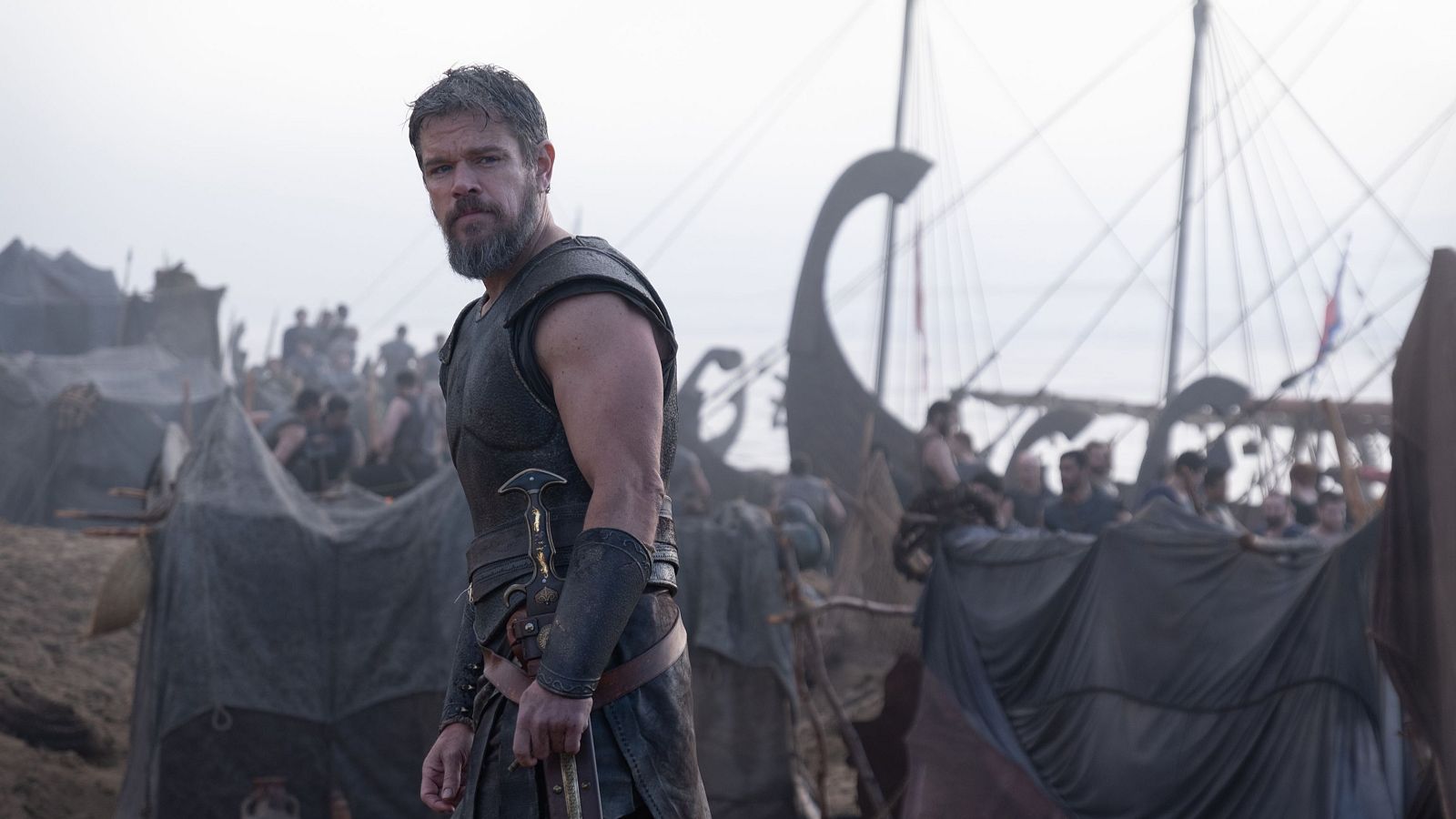 Matt Damon con armadura y barba canosa, en un campamento con barcos antiguos.