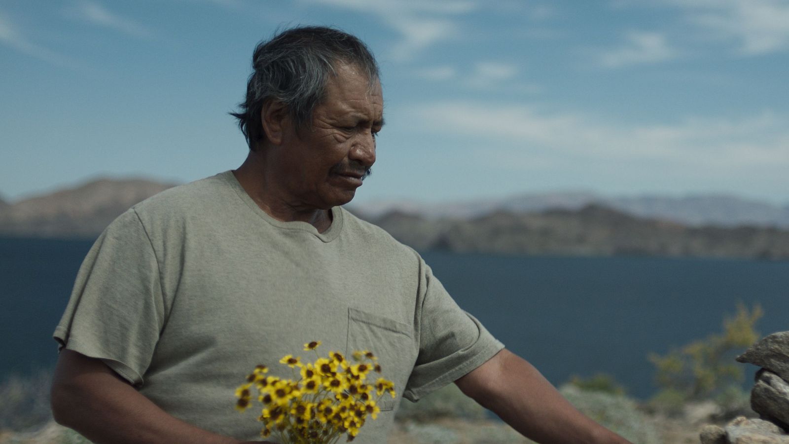 Hombre mayor con flores amarillas en entorno natural, junto a agua y montañas.