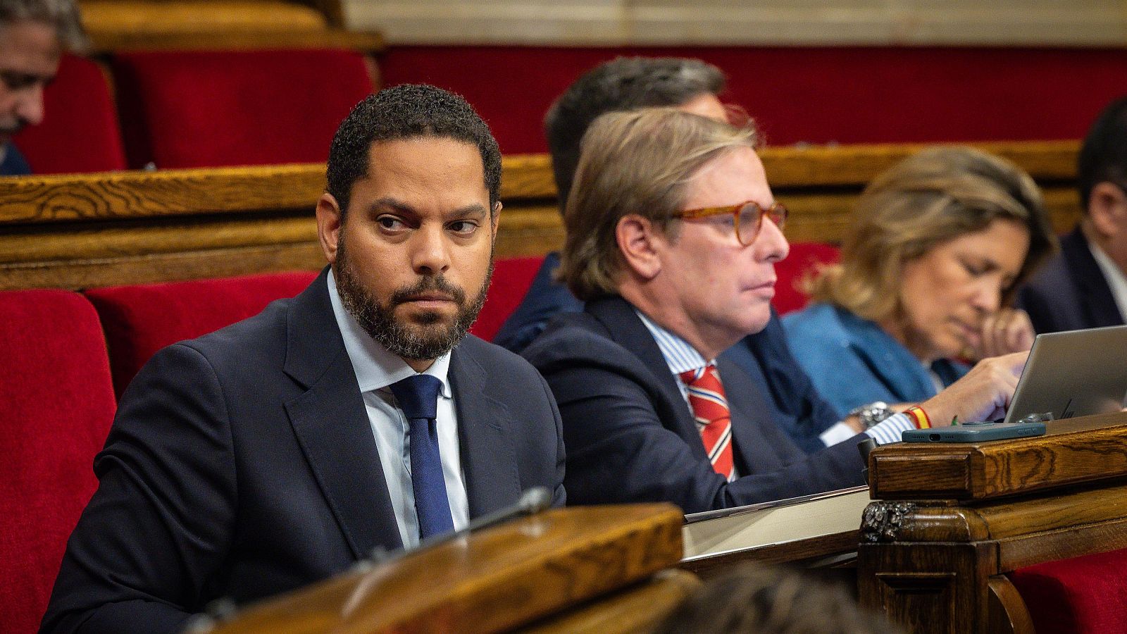 Ignacio Garriga y Joan Garriga de Vox en el Parlament. Ignacio con traje oscuro y corbata azul, Joan con gafas naranjas y traje a rayas. Una mujer con pulsera española mira un dispositivo.