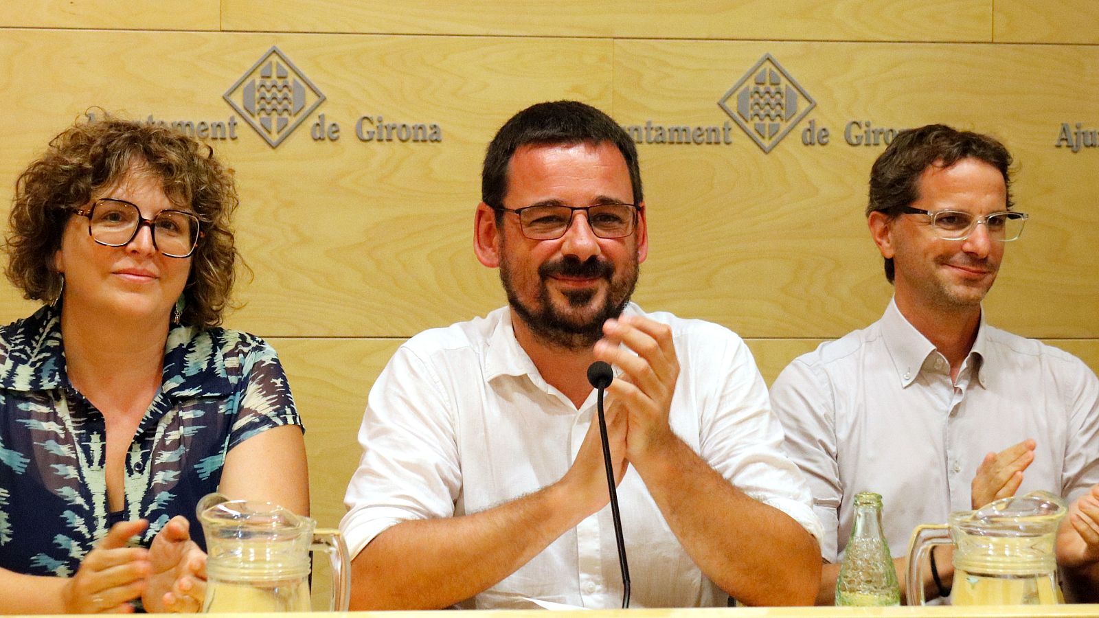 Lluc Salellas, alcalde de Girona, aplaude junto a dos personas en un evento del Ayuntamiento. El fondo muestra el escudo y el nombre de la ciudad.