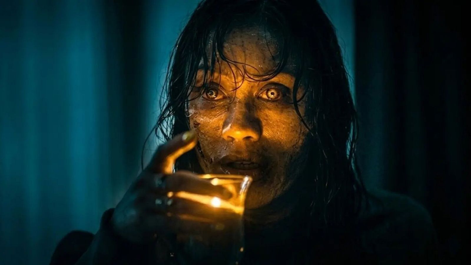 Primer plano aterrador de una mujer con ojos claros y dilatados, sosteniendo un vaso con luz naranja que ilumina su rostro de forma inquietante.