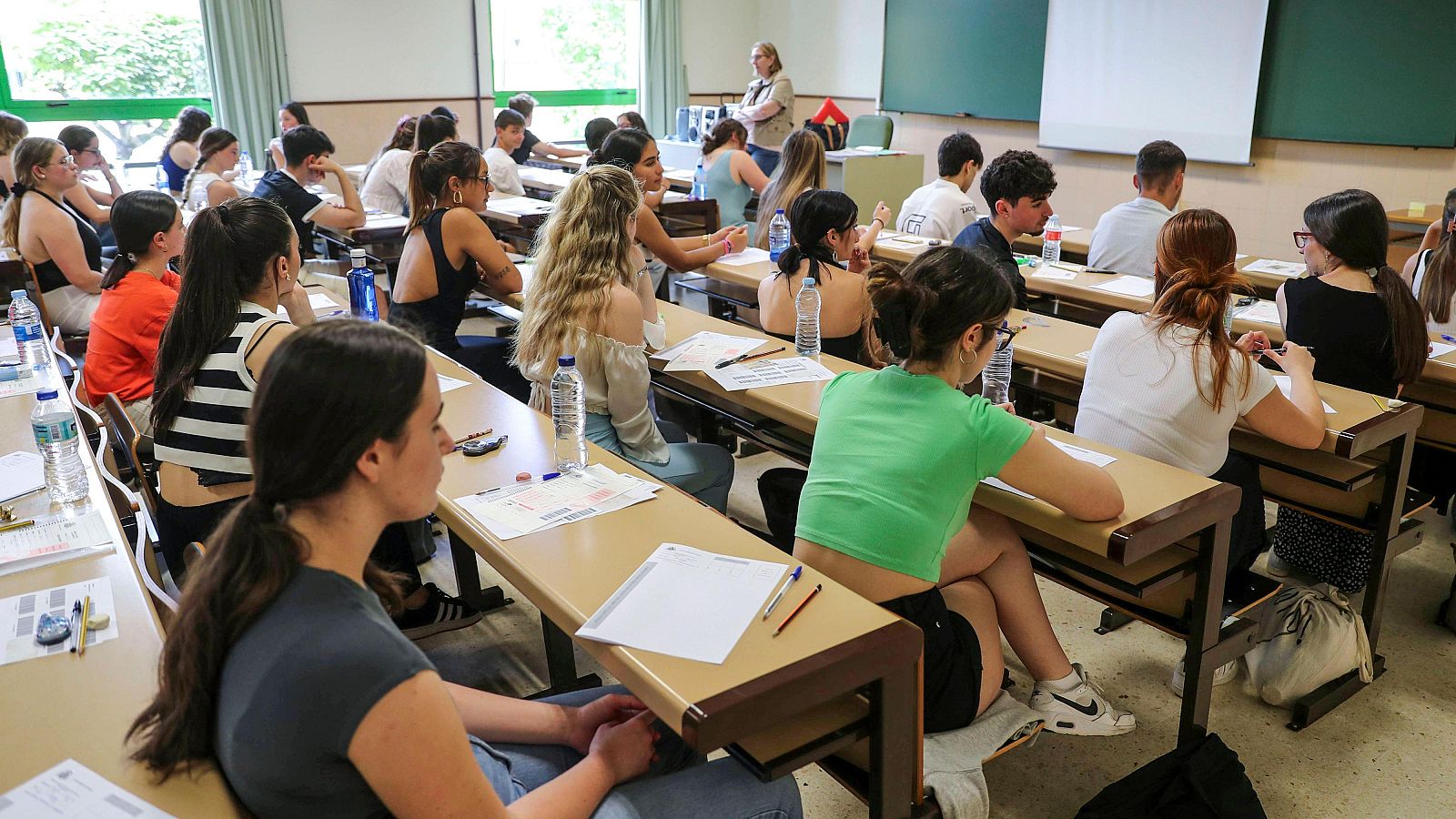 Estudiantes realizando un examen en un aula, con mesas individuales, materiales de escritura y un supervisor presente.