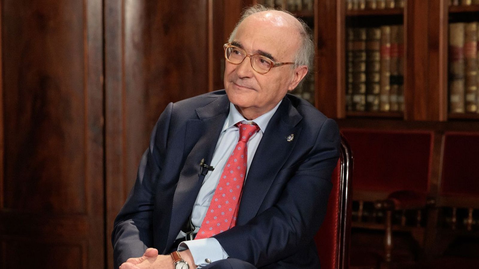 Retrato de Benigno Pendás con traje azul, camisa clara y corbata roja, en un entorno de estudio con estanterías de libros y sillas de terciopelo.