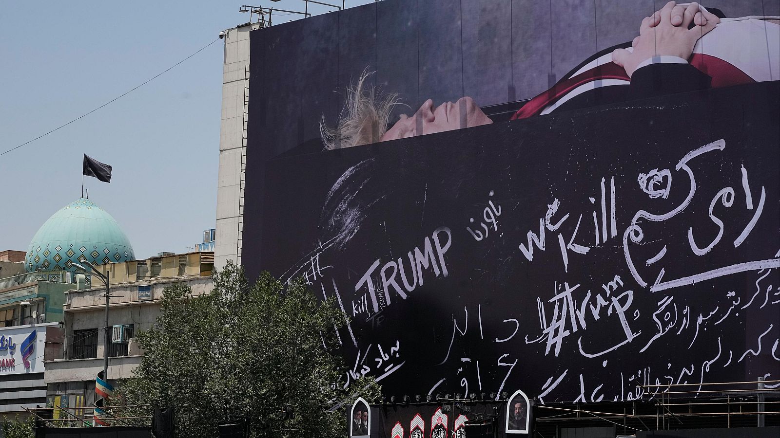 Valla publicitaria en Teherán con Donald Trump en un ataúd y mensajes anti-Trump en inglés y persa. Se observa una cúpula turquesa y un árbol.