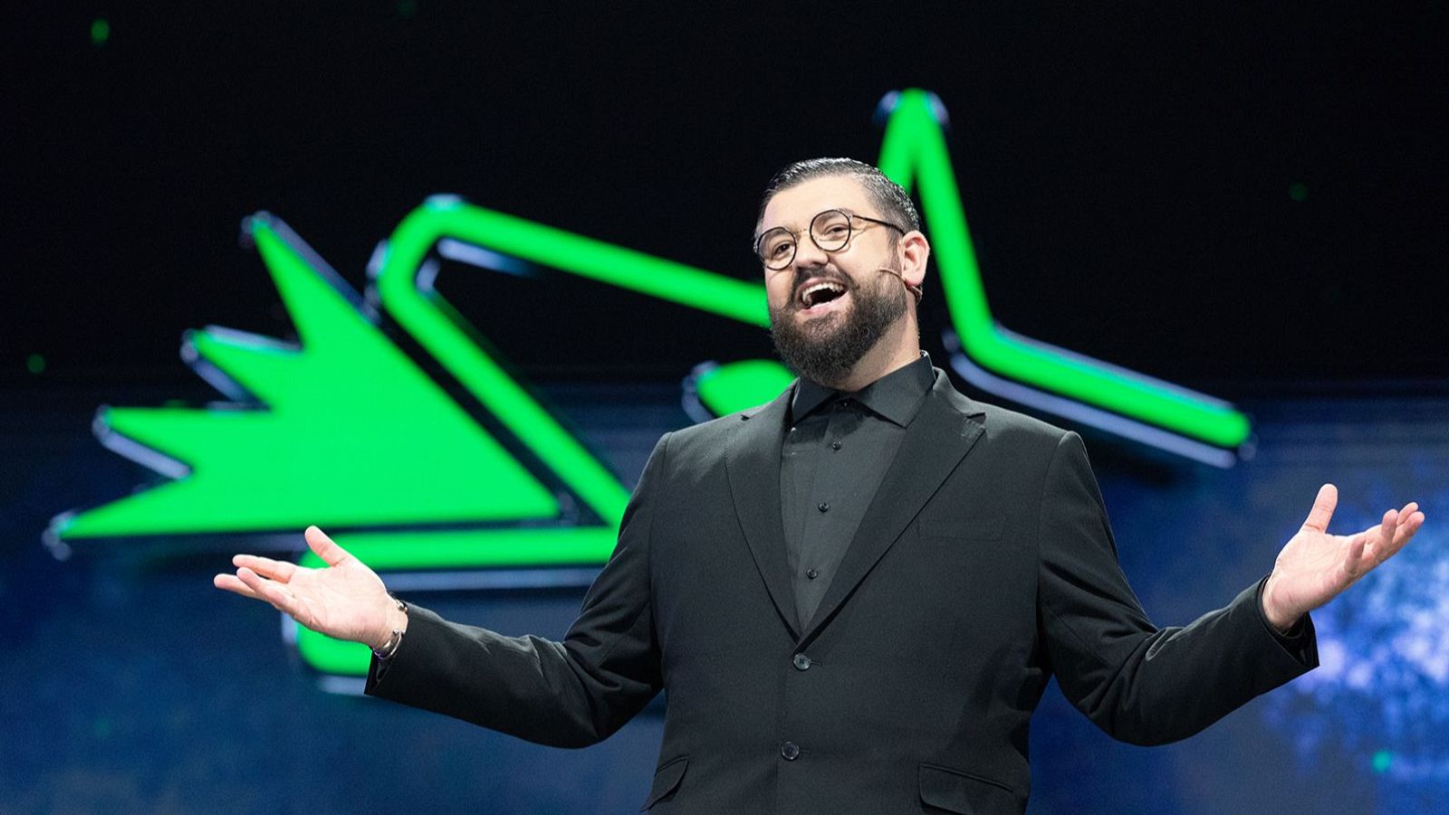 Manu Sánchez, presentador con traje oscuro y gafas, sonríe ampliamente en un escenario con gráfica verde neón y fondo oscuro. Sus brazos están extendidos en un gesto acogedor.