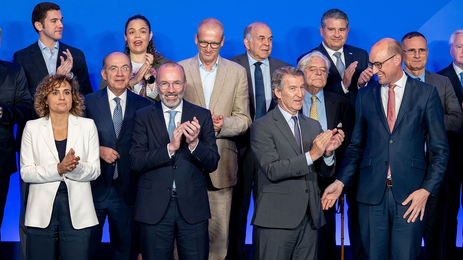 Grupo de personas, incluyendo a Cuca Gamarra, Alberto Núñez Feijóo y Manfred Weber, aplaudiendo y sonriendo en un evento con fondo azul.