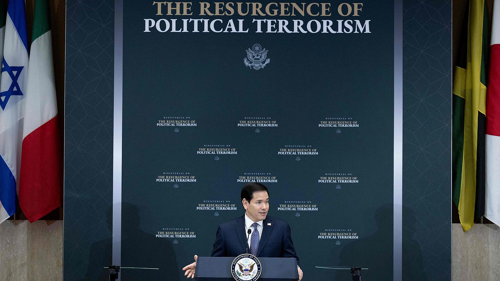 Marco Rubio habla en un evento con el titular "THE RESURGENCE OF POLITICAL TERRORISM" y el Gran Sello de EE.UU. de fondo. Banderas de Israel, Italia, Jamaica y Canadá flanquean la escena.
