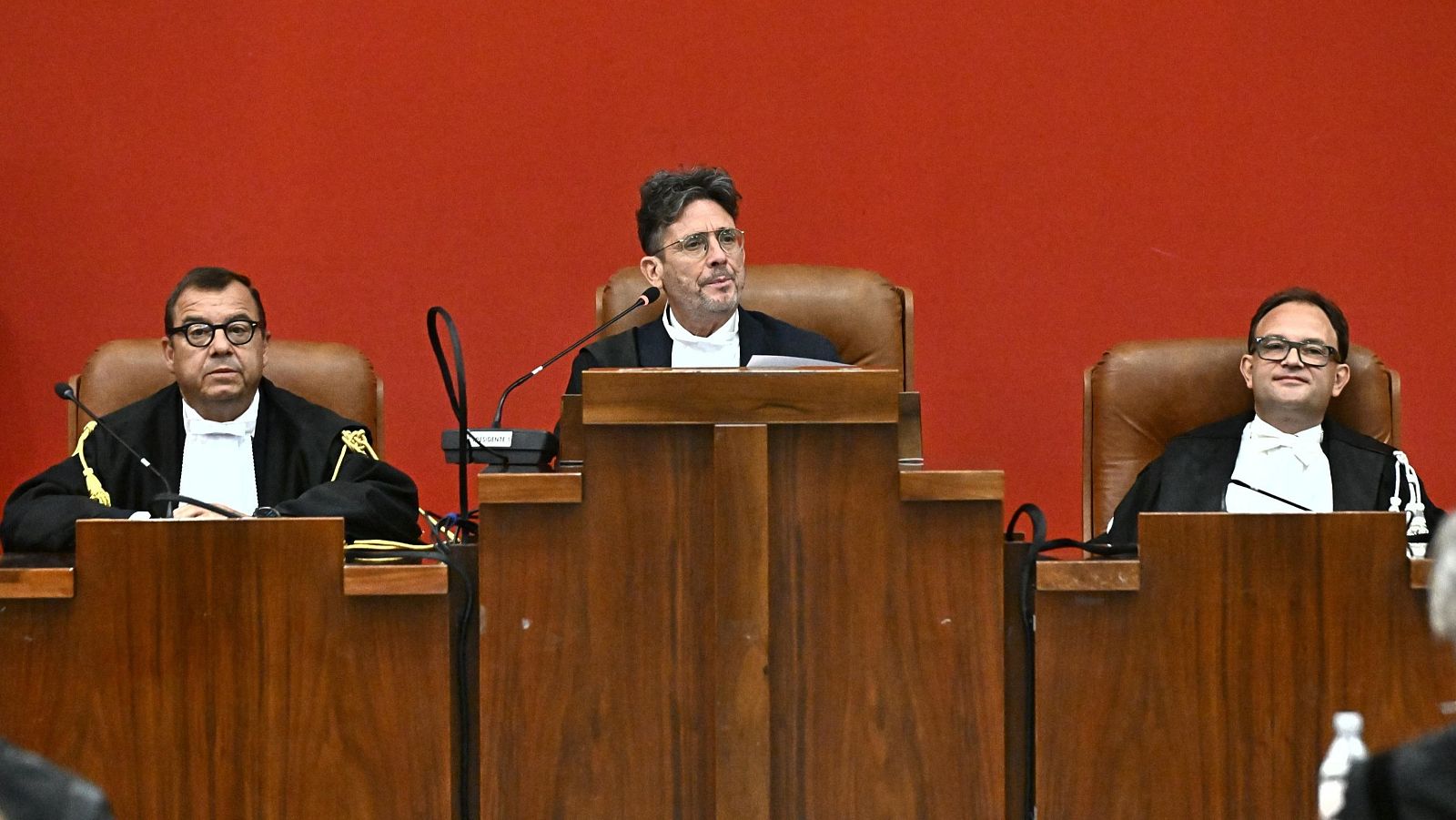 Panel de tres jueces con togas negras y gafas, sentados en un tribunal con fondo rojo, listos para emitir una sentencia.