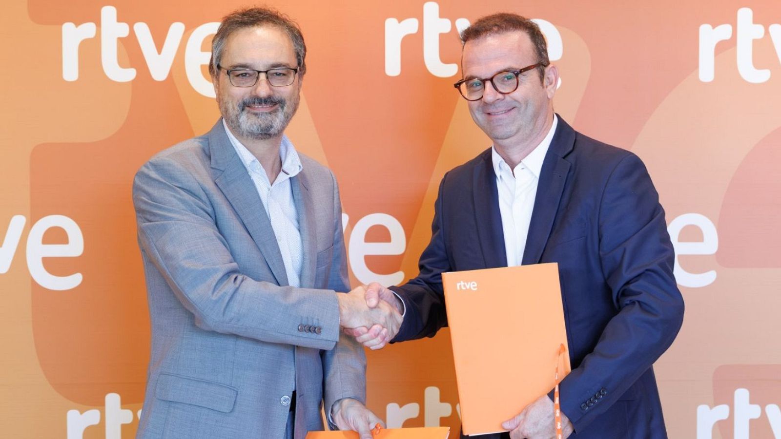 Alfonso Morales y Roberto Suárez Martín, con gafas y trajes, se estrechan la mano frente a un fondo naranja con el logo de RTVE. Ambos sostienen carpetas naranjas.