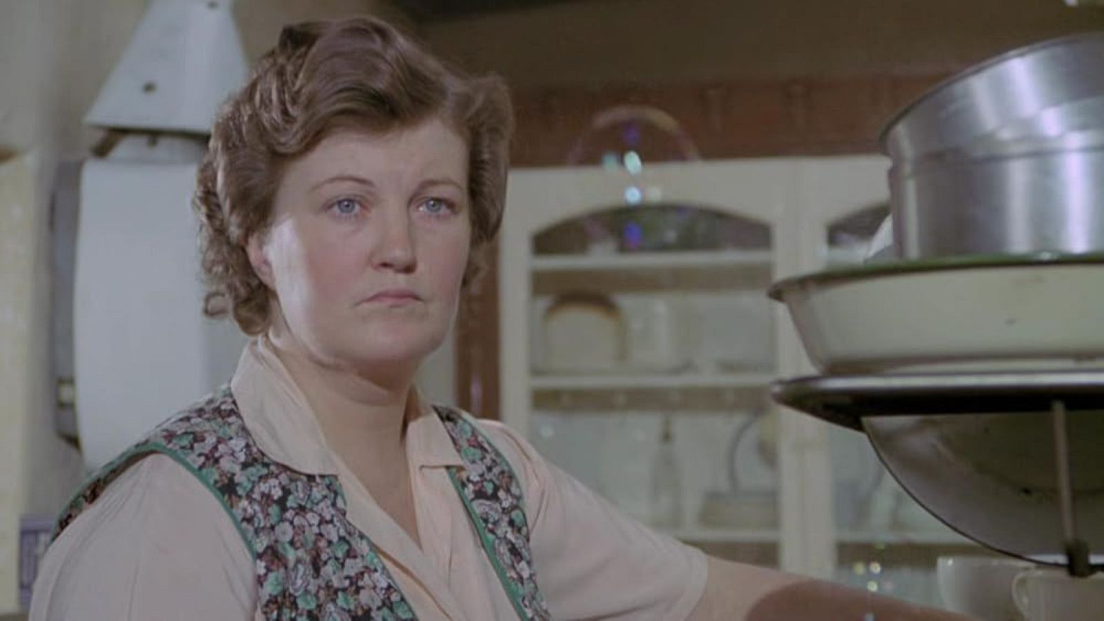 Primer plano de Brenda Fricker con blusa y delantal floral, en un entorno de cocina con utensilios apilados. Expresión seria y ojos azules intensos.