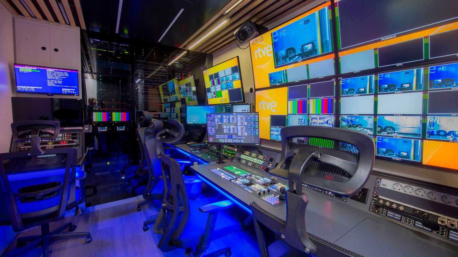 Sala de control de unidad móvil de RTVE con muro de pantallas mostrando el logo de RTVE, barras de color y textos como POMMIX y TEA. Puestos de trabajo con paneles de control y sillas iluminadas.