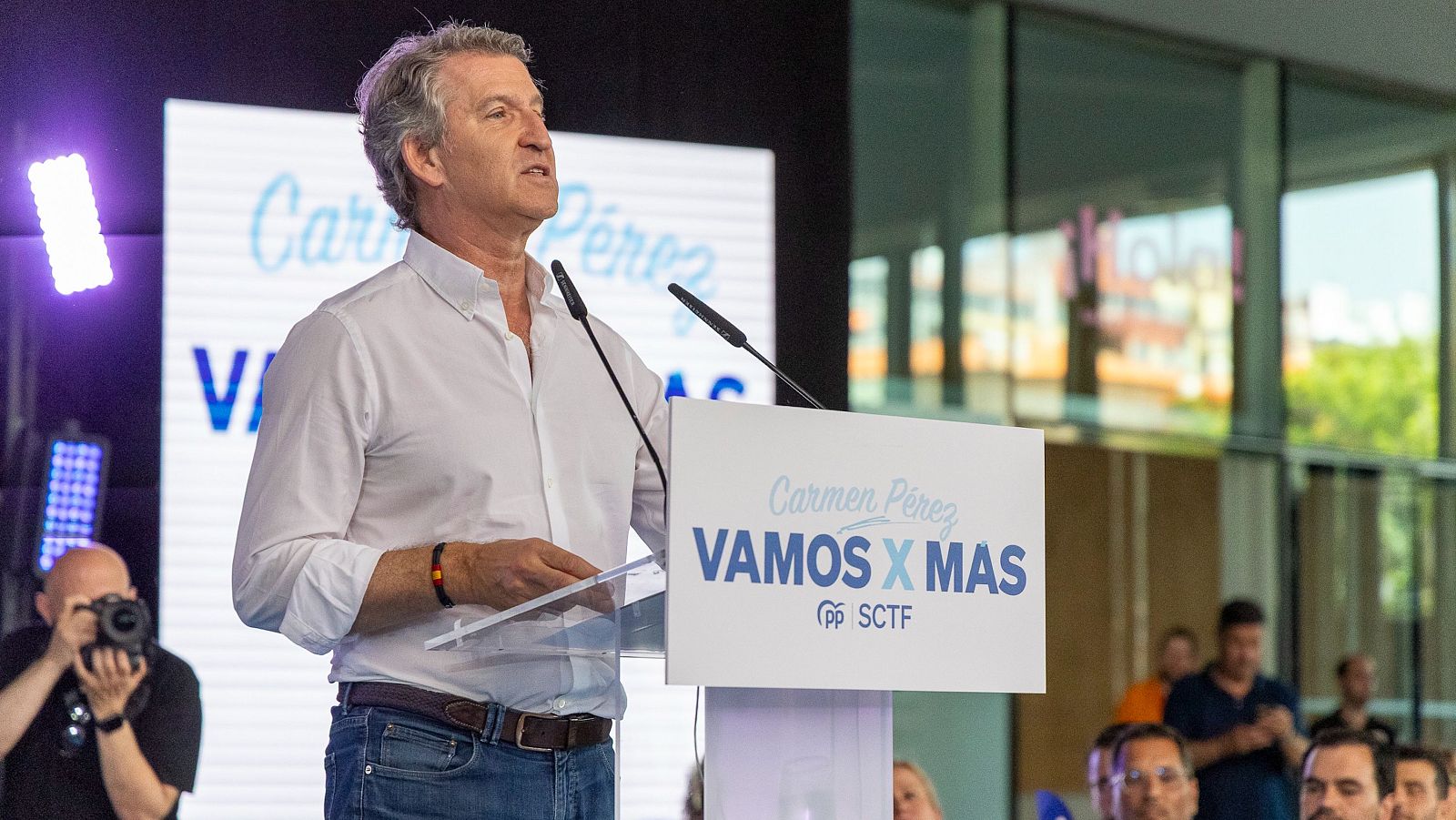 Alberto Núñez Feijóo habla en un evento político. En el podio y pantalla se lee "Carmen Pérez" y "VAMOS X MAS". Un fotógrafo documenta la escena.
