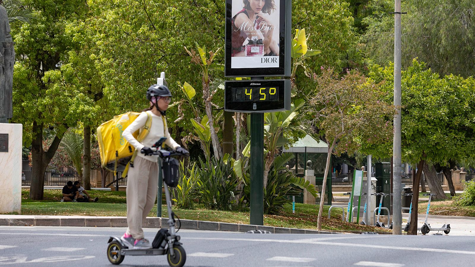 Una repartidora con casco y mochila amarilla conduce un patinete eléctrico bajo un sol abrasador, mientras un termómetro marca 45°C junto a un anuncio de "Miss Dior".
