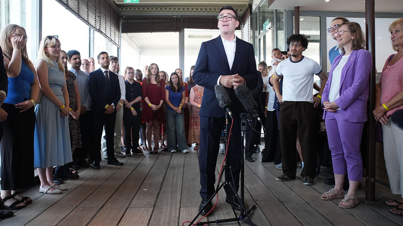 Andy Burnham, líder del Partido Laborista, habla en un evento público rodeado de asistentes con pulseras amarillas, en un espacio interior luminoso con suelo de madera.