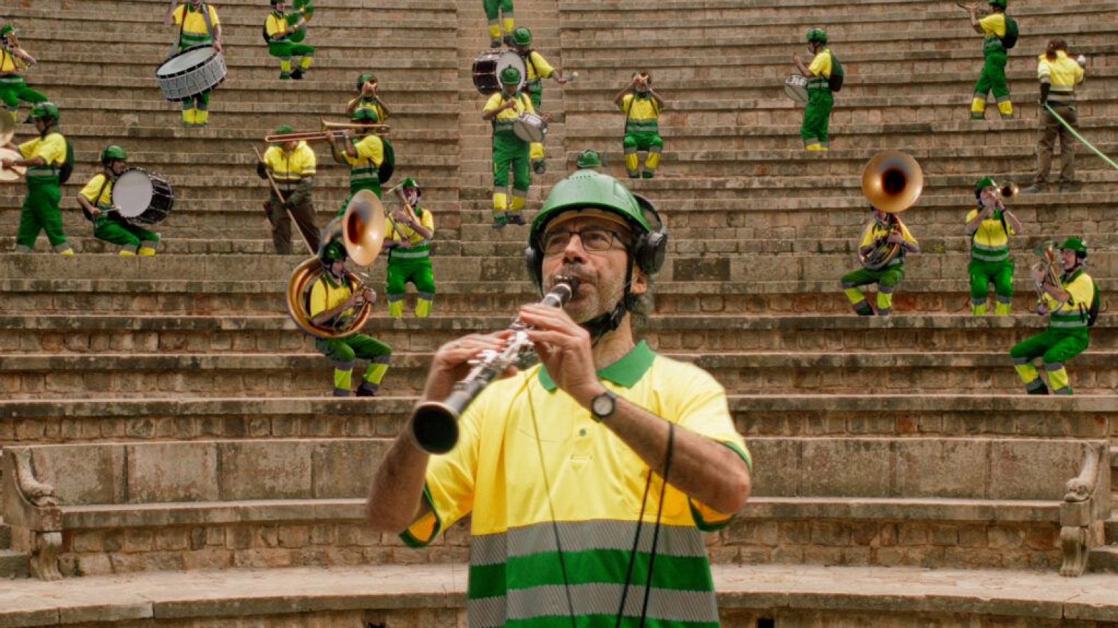 Músico con casco verde y camiseta amarilla/verde toca clarinete, rodeado por una orquesta en gradas de piedra de anfiteatro.