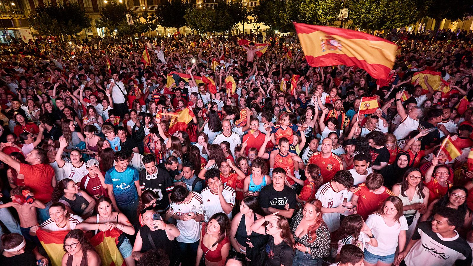 Multitud celebra con banderas de España y euforia en un evento nocturno al aire libre. Rostros llenos de alegría y orgullo colectivo.