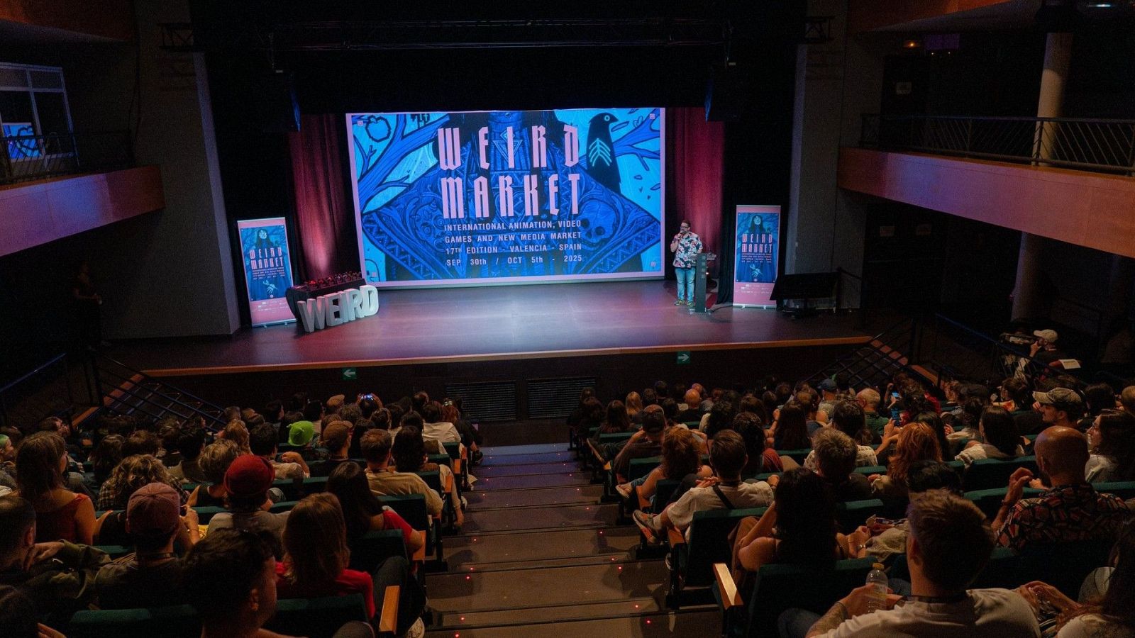 Escena de un auditorio con el escenario iluminado, mostrando la pantalla principal del "WEIRD MARKET" (13ª edición, Valencia, España, 28 Sep - 3 Oct 2023) y un ponente. El público asiste al evento.