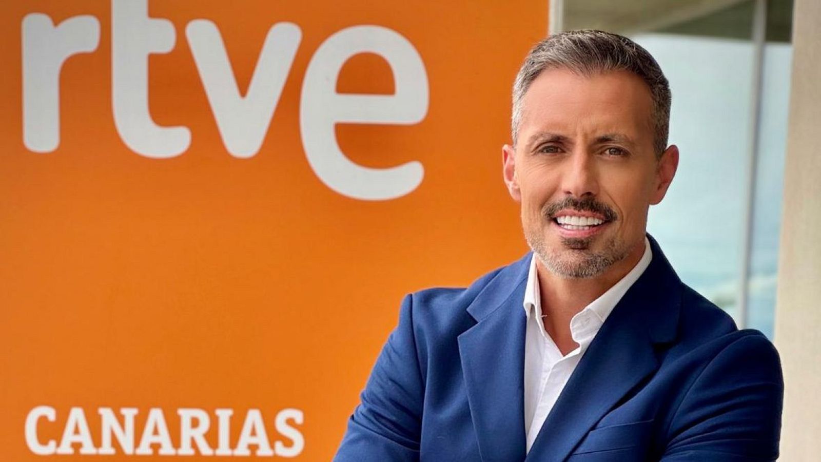 Periodista español Miguel Ángel Guerra sonríe a cámara con traje azul marino, camisa blanca y fondo naranja con logo RTVE y "CANARIAS".