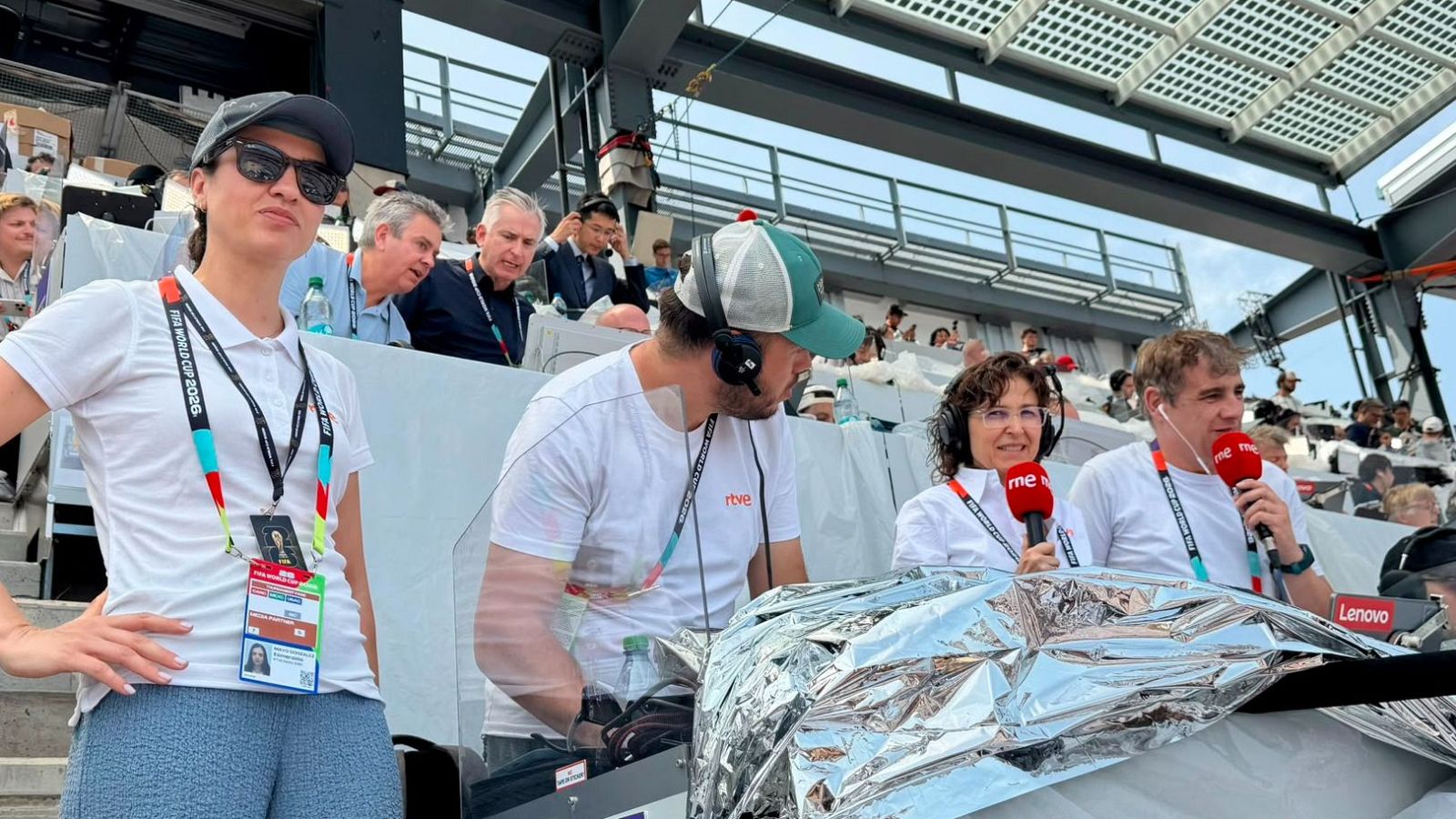 Periodistas de RNE en una cabina de retransmisión, con acreditaciones de la FIFA WORLD CUP 2026 y logos de rtve y rne visibles. Se observa equipo técnico y gradas del estadio al fondo.