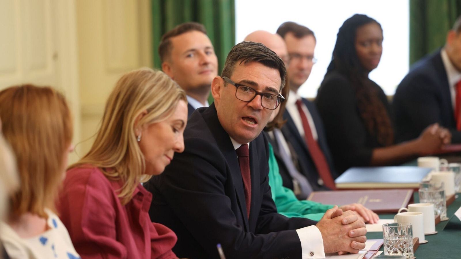 Andy Burnham, con gafas y traje, lidera una reunión formal. Participan otras personas, algunas observando atentamente. La mesa tiene documentos y vasos.