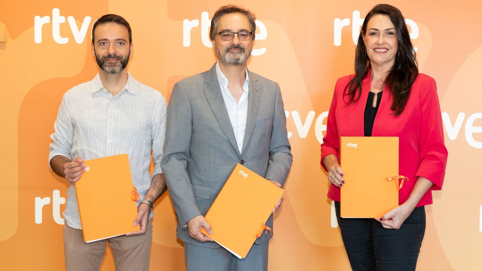 Iván Agenjo, Alfonso Morales y Nathalie Martínez posan sonrientes con carpetas de RTVE frente a un fondo naranja con el logo de la cadena.