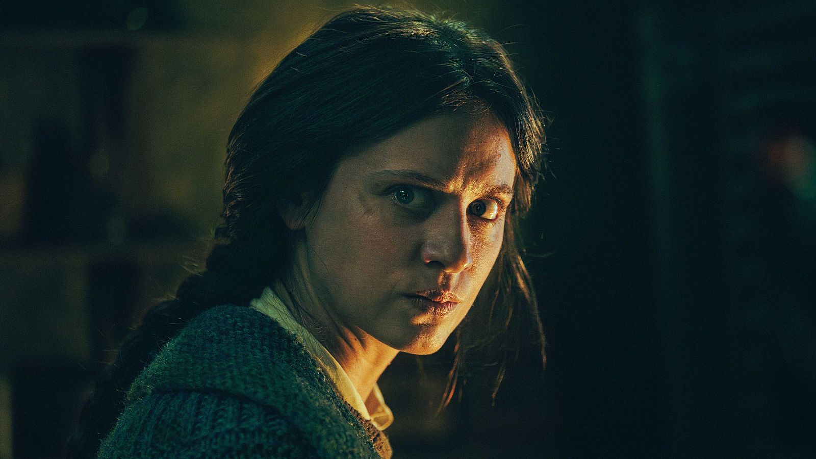 Primer plano de Michelle Jenner con expresión seria y cabello oscuro trenzado, en "El nido". Iluminación dramática y fondo oscuro acentúan su rostro.