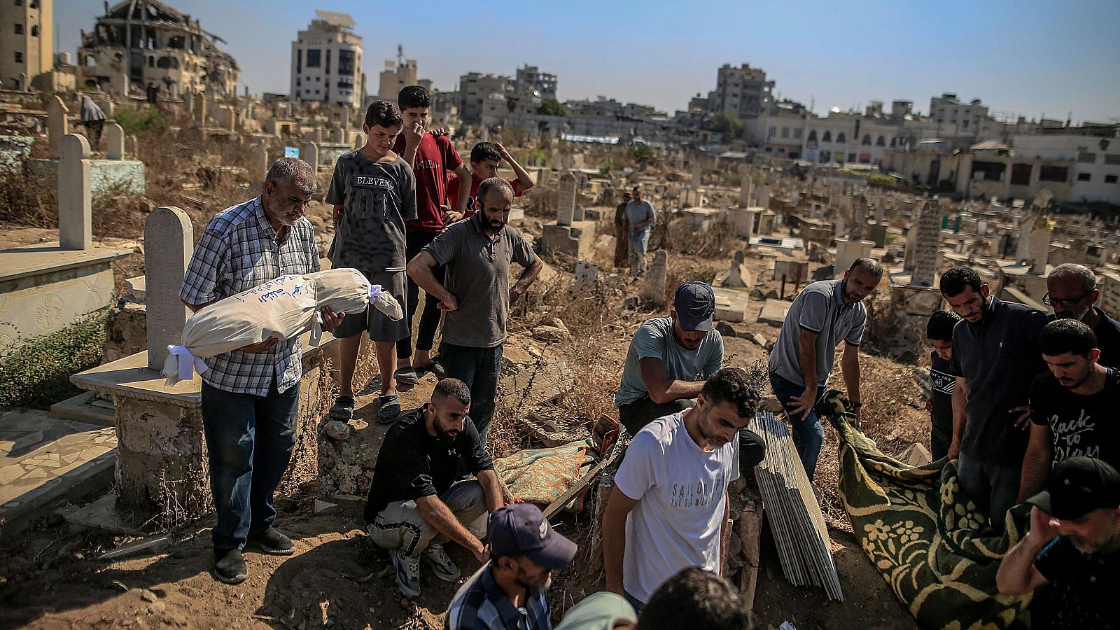 Ceremonia de entierro palestina en un cementerio. Hombres y jóvenes participan, uno sostiene un bulto envuelto en tela blanca. Al fondo, edificios dañados sugieren un conflicto.