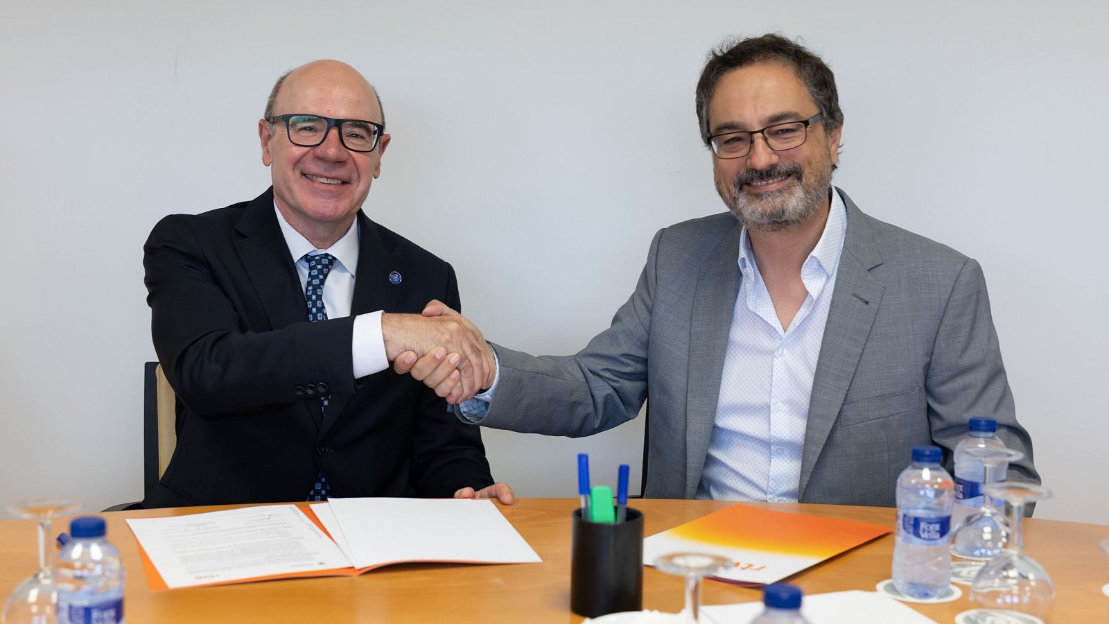 Pedro Mercado, rector de la UGR, y Alfonso Morales, secretario general de RTVE, sonríen mientras se estrechan la mano sobre una mesa con documentos y botellas de agua.